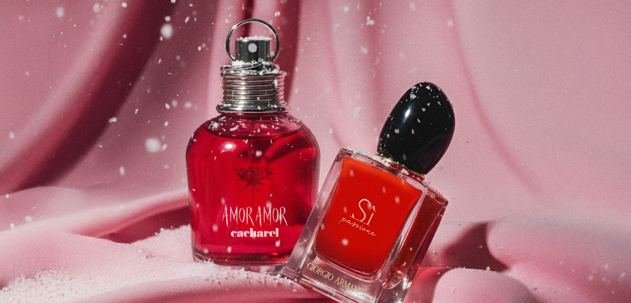 Due bottiglie di profumo sono posizionate su uno sfondo di satin rosa. La bottiglia di Cacharel è in vetro rosso con un collo in argento e un design a rosa. La bottiglia di Giorgio Armani è quadrata, con un liquido di un rosso brillante e un tappo rotondo nero.