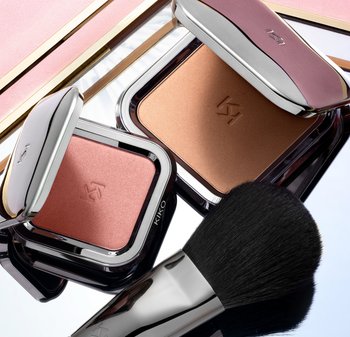 Two square compacts of blush, one pink and one bronze, with silver accents, alongside a soft black makeup brush on a reflective surface.
