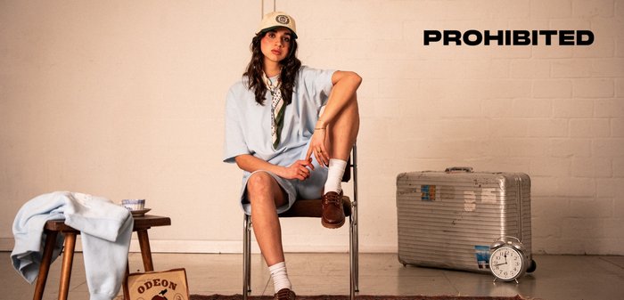 Light blue oversized shirt, white socks, brown shoes. Silver suitcase and clock on the floor. "PROHIBITED" text in black.