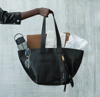Mano che tiene una borsa tote in pelle nera contenente una bottiglia d'acqua, occhiali da sole, buste, fazzoletti e un quaderno marrone davanti a un muro grigio.