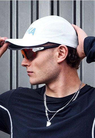 Man wearing white cap, dark sports sunglasses, black athletic top, and silver necklaces, adjusting cap with hand behind head against gray background.