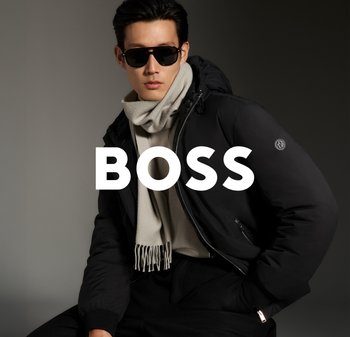 Zwarte gewatteerde jas met capuchon, grijze sjaal met franjes en zwarte zonnebril. Model poseert tegen een donkere achtergrond. Gedurfde tekst "BOSS" in het wit.