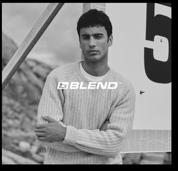 Lichtgrijze gebreide trui met een gestructureerd patroon, gedragen door een man die met gekruiste armen voor een groot bord staat. Zwart-wit foto.