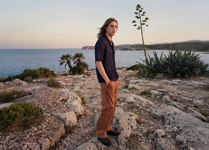 Jeune homme aux longs cheveux se tient sur une côte rocheuse près de la mer, portant une chemise foncée et un pantalon marron au coucher du soleil.