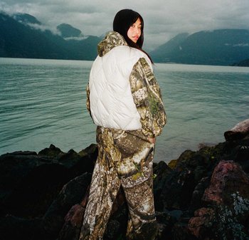 Donna con giacca mimetica e pantaloni, con gilet imbottito bianco, in piedi su una riva rocciosa, di fronte a un lago con montagne nebbiose sullo sfondo.