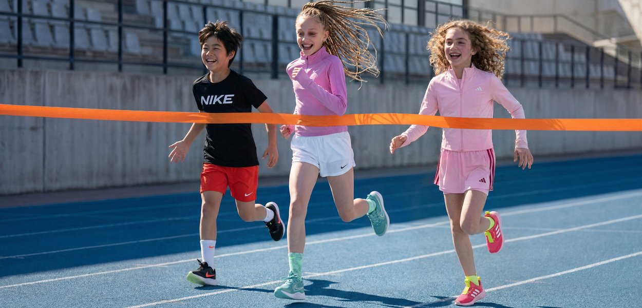 Three children joyfully sprinting on a blue track, crossing the orange finish line tape during a race.