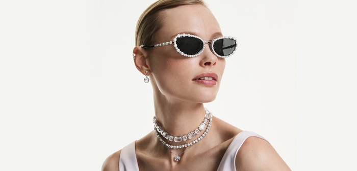 Woman wearing white sleeveless top, black oval sunglasses with rhinestone frame, layered crystal necklaces, and crystal drop earrings.