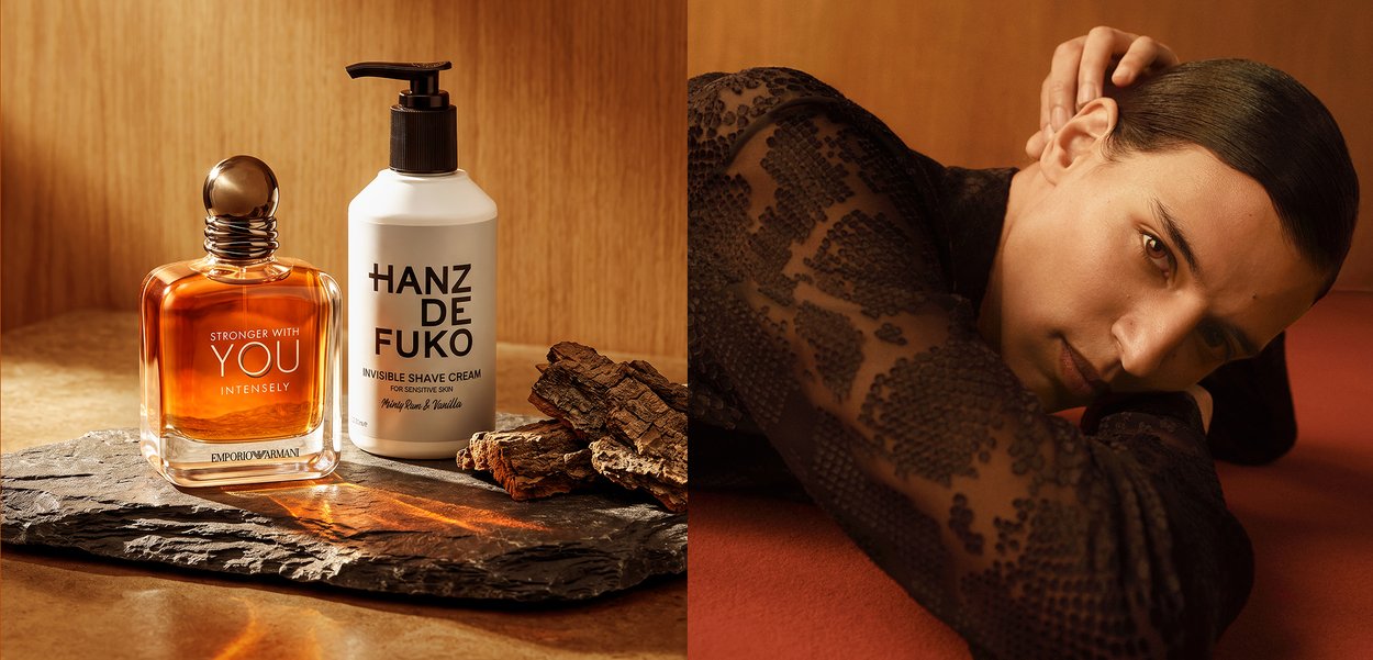 Cologne and shave cream on stone surface beside a man in a lace top leaning on his arm against a brown background.
