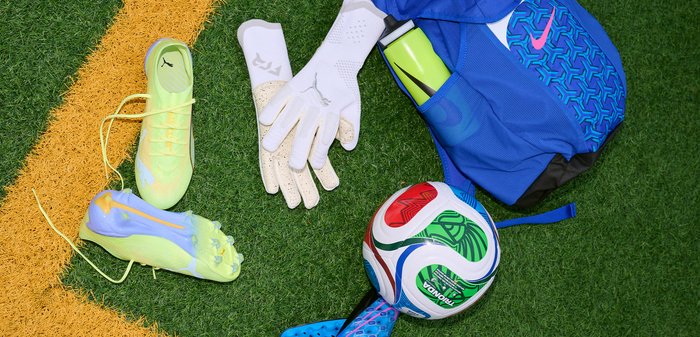 Κίτρινα ποδοσφαιρικά παπούτσια Puma, λευκά γάντια τερματοφύλακα, μπλε αθλητική ενδυμασία με ροζ λογότυπο Nike, πράσινο μπουκάλι νερού και πολύχρωμη ποδοσφαιρική μπάλα σε χλοοτάπητα κοντά σε κίτρινη γραμμή.