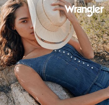 Kvinne som ligger tilbakelent på steiner iført en ermeløs denimtopp, holder en vevd hatt som delvis dekker ansiktet hennes utendørs med Wrangler-logo over.