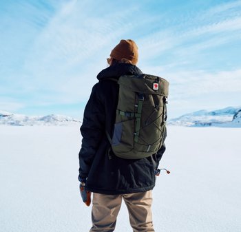 Grüner Rucksack mit Roll-Top-Design, verstellbaren Trägern und grauen Akzenten, getragen über einer schwarzen Jacke und beigen Hosen in einer schneebedeckten Landschaft.