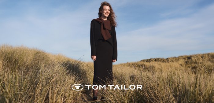 Lachende vrouw in een zwarte lange jurk en een bruine sjaal staat in het hoge gras onder een blauwe lucht op een strand of duingebied.