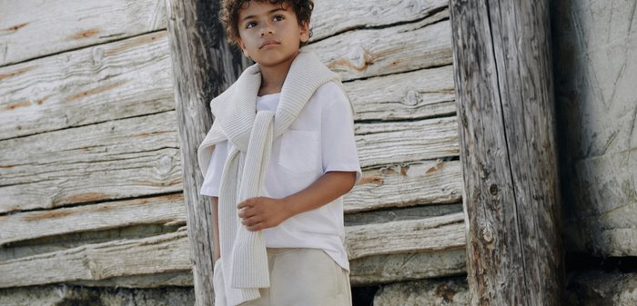 Jeune garçon aux cheveux bouclés debout pieds nus sur le sable, vêtu d'un short beige, d'une chemise blanche et d'un pull posé sur les épaules, près d'un mur en bois usé par le temps.