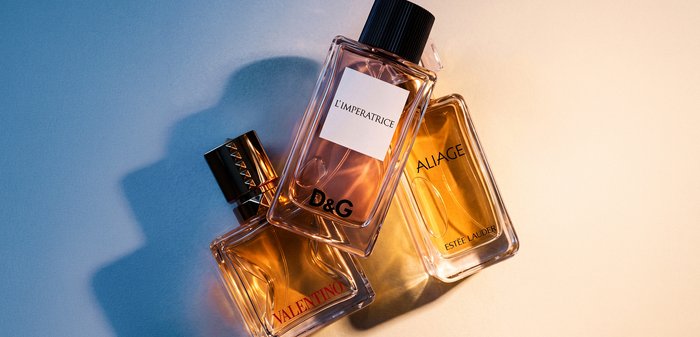 Trois bouteilles de parfum rectangulaires étiquetées Valentino, D&G L'Imperatrice et Estée Lauder Aliage sur une surface bleue et beige.