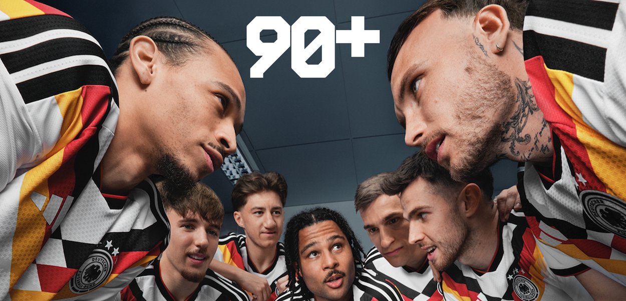 Sieben männliche Fußballspieler, die Deutschland-Trikots tragen, stehen eng zusammen und konzentrieren sich intensiv während eines Spiels, das bei 90+ Minuten pausiert wurde.