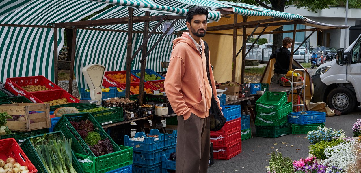 Un hanorac piersiciu, pantaloni gri și o geantă din plasă purtate de o persoană într-un market, cu lăzi colorate de fructe și legume sub un cort cu dungi.