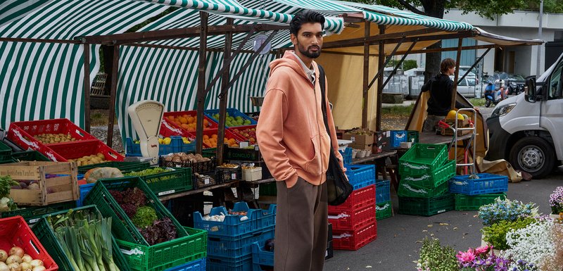 Sweatshirt pêche, pantalon gris et sac en filet portés par une personne dans un marché avec des caisses colorées de fruits et légumes sous une canopy rayée.