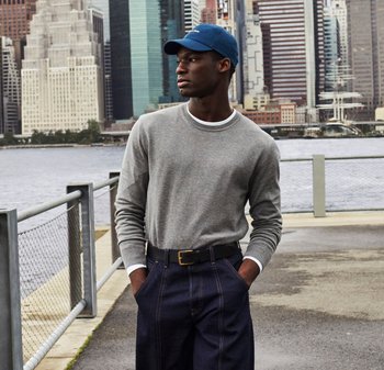 Grijze lange mouwen shirt met witte halslijn, donkerblauwe pet en marineblauwe brede broek. Staan bij water met een stedelijke skyline op de achtergrond.