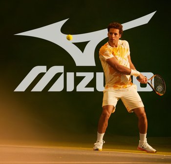 Jugador de tenis con una camiseta amarilla y blanca de patrón geométrico y pantalones cortos blancos, sosteniendo una raqueta, con una pelota de tenis amarilla en movimiento.