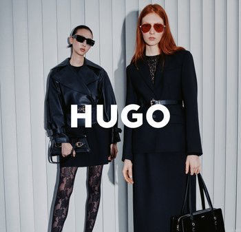 Twee modellen staan tegen verticale jaloezieën. De een draagt een zwart leren jas en kanten panty, de ander in een donker kostuum met een riem en handtas.