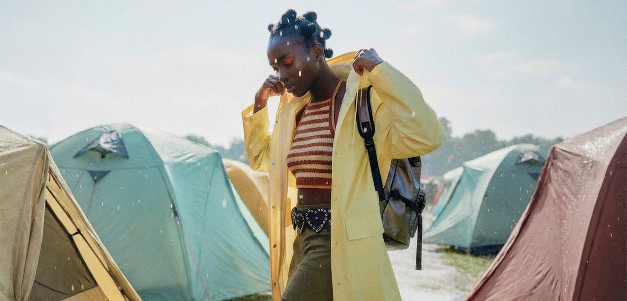 Joven mujer con camiseta de rayas y chubasquero amarillo ajustándose el abrigo cerca de tiendas de campaña en un área de camping al aire libre bajo la lluvia.