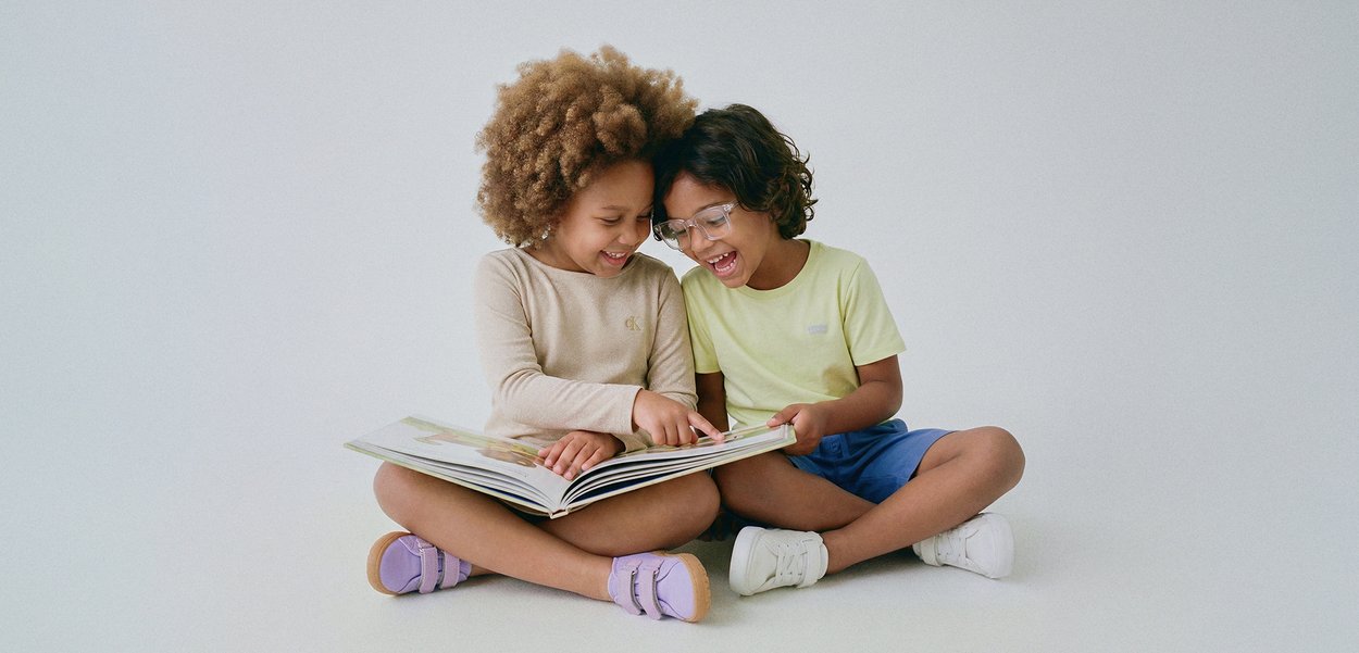 Dos niños sentados con las piernas cruzadas, sonriendo y leyendo juntos un libro grande abierto sobre un fondo sencillo.