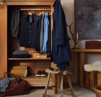 Houten kast met een marineblauwe jas, een doorgestikte vest, diverse hemden en gevouwen kledingstukken. Accessoires zijn onder andere een bruine tas, bruine schoenen en een zonnebril.