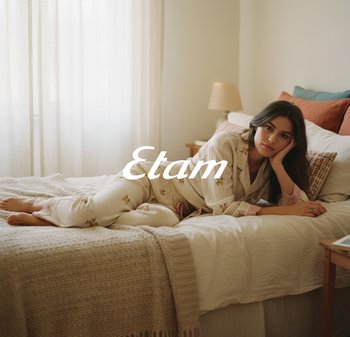 Woven, cremefarbene Pyjamas mit floralen Stickereien, die einen Kragen und rosa Akzente aufweisen. Eine strukturierte Decke liegt auf dem Bett. Sanfte Beleuchtung.