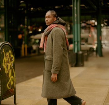 Grijze wollen jas met grote zak, gedragen over een zwarte hoodie, aangevuld met een kleurrijke sjaal. Stedelijke achtergrond.