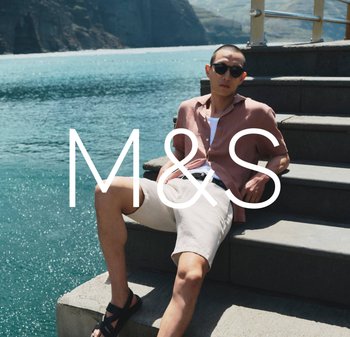 Man wearing sunglasses, short-sleeve shirt, and shorts reclining on concrete steps by blue water with rocky cliffs in the background.