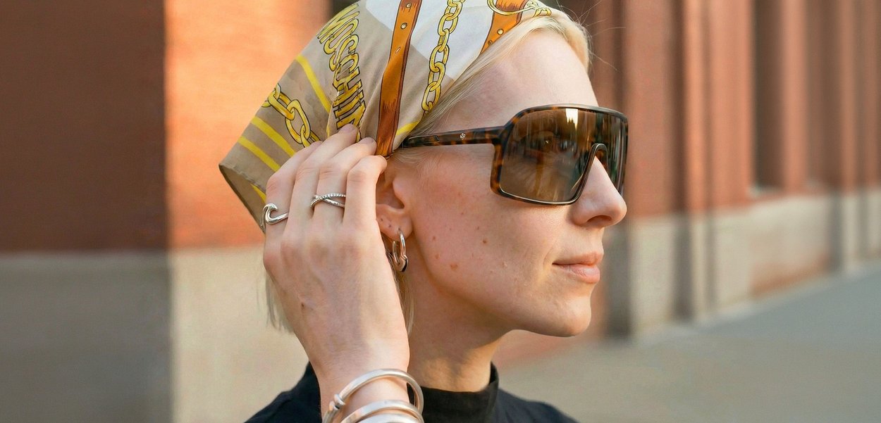 Mujer con cabello rubio que lleva gafas de sol carey y una bufanda de seda con estampado, ajustando la bufanda al aire libre cerca de un edificio de ladrillo.