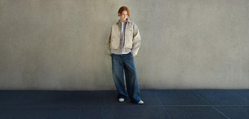 Personne avec des cheveux roux portant une veste beige, des hauts gris superposés, un jean large bleu et des chaussures blanches se tient sur un sol à motif de grille sombre contre un mur gris.