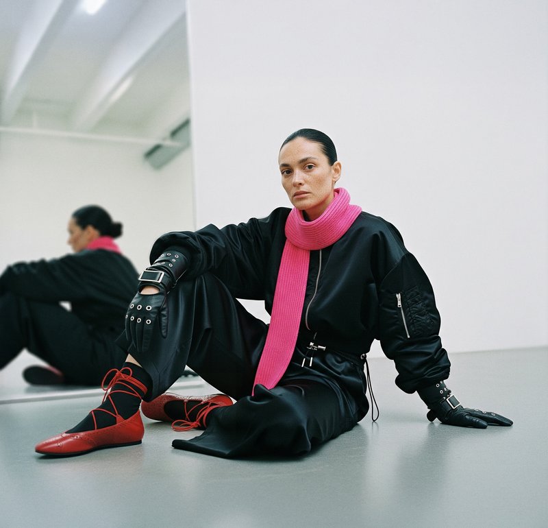 Black oversized jacket, pink knitted scarf, black gloves with silver hardware, red ballet flats with crisscross laces, grey floor, mirrored background.