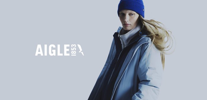 Doudoune bleu clair avec fermeture éclair sur le devant, superposée à un pull en polaire blanc, associée à un bonnet bleu à côtes. Nom de la marque "AIGLE 1853" affiché.