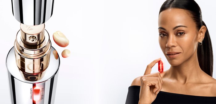 Primo piano di una bottiglia cosmetica con pompa lucida argento e oro che eroga gocce di crema liquida beige su sfondo bianco.