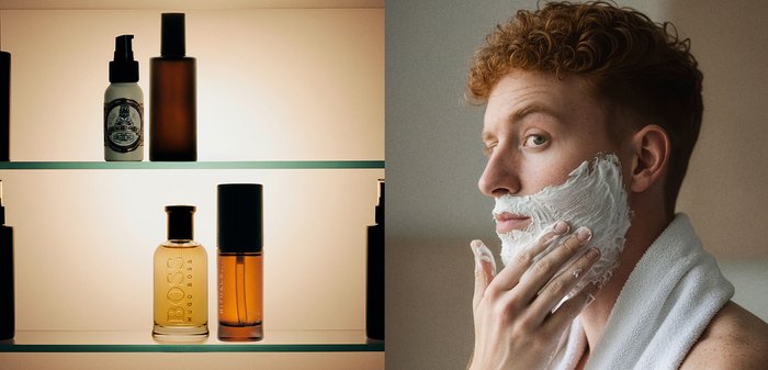 Uomo con un asciugamano sulle spalle applica crema da barba sul viso accanto a scaffali che contengono bottiglie di prodotti per la cura personale e della pelle.