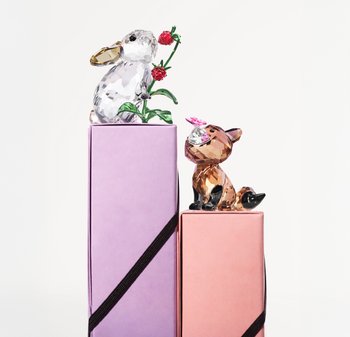 Ein kristallener Hase mit einer Blume sitzt auf einer lila Box, während ein brauner Kristallfuchs mit einer rosa Blume auf einer rosa Box ruht.