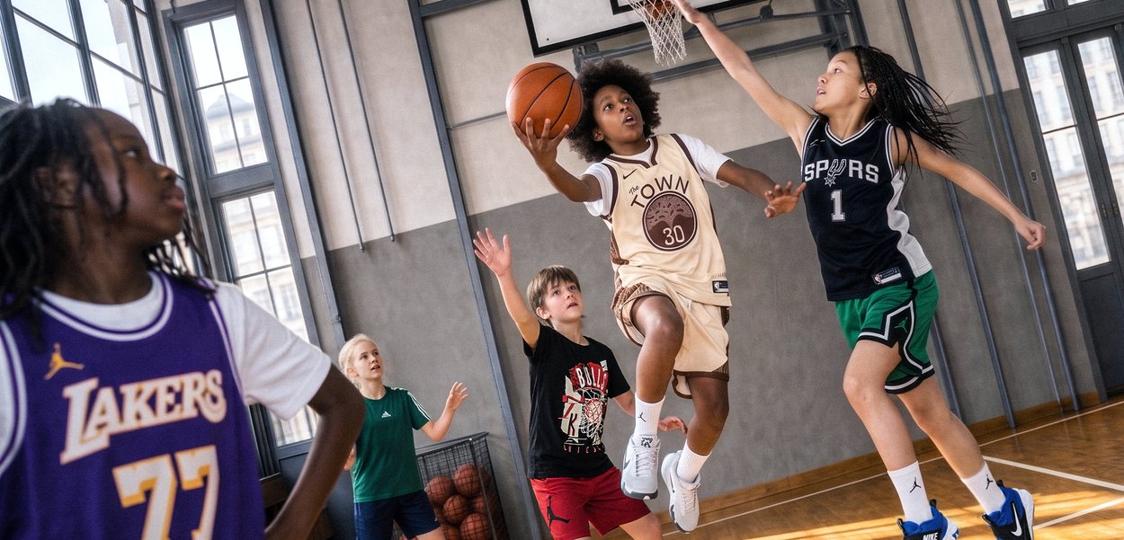 Children playing basketball indoors, one jumping to shoot while others defend and watch on a polished wooden court with large windows.