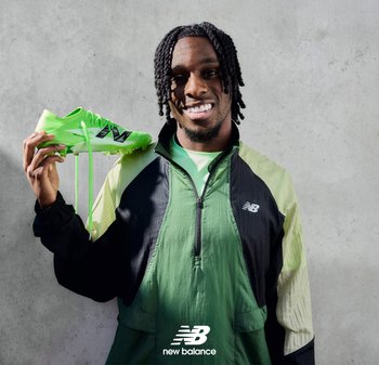 Scarpa da calcio sintetica verde con accenti neri, tenuta da una persona in un giubbotto a due tonalità verde e nero, sullo sfondo di un murale di cemento.