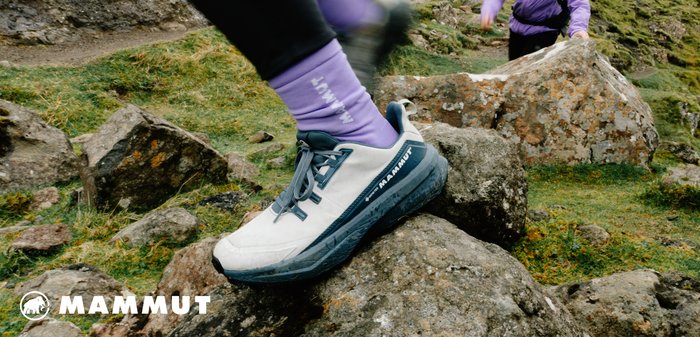 Hellgraue Wanderschuhe mit blauen Akzenten, ausgestattet mit einer strukturierten Sohle, Schnürsenkeln und Logo. Getragen mit lila Socken auf steinigem Terrain.