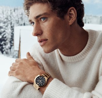 Reloj de oro con esfera negra, con números romanos y una pulsera metálica. Modelo con suéter crema, con un fondo nevado.