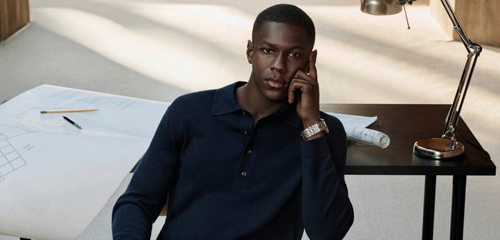 Joven con camisa oscura sentado pensativamente en un escritorio con planos arquitectónicos, lápiz, bolígrafo y lámpara de escritorio bajo una suave luz natural.