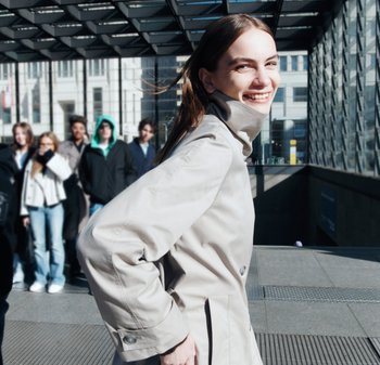 Helle beige Trenchcoat mit hohem Kragen, glatter Oberfläche und geknöpftem Front. Das Modell steht im Sonnenlicht, lächelnd, mit urbanem Hintergrund.