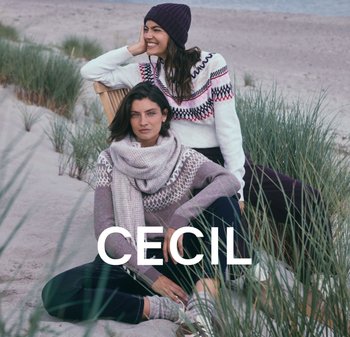 Zwei Frauen sitzen am Strand und tragen gemusterte Strickpullover. Die eine trägt einen weißen Pullover mit pinken und schwarzen Designs, die andere einen grauen mit weißen Akzenten.