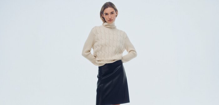 Woman wearing a cream cable knit turtleneck sweater, black leather skirt, and black knee-high boots standing against a plain background.