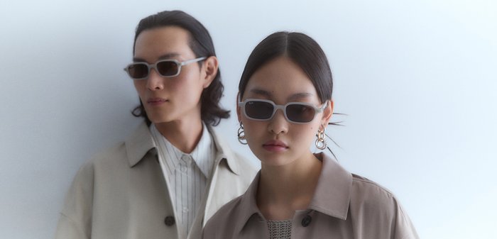 Okulary przeciwsłoneczne z szarym ramkiem i ciemnymi soczewkami, w eleganckim prostokątnym kształcie. Modelka ma na sobie jasnobrązowy płaszcz i duże kolczyki w kształcie koła.