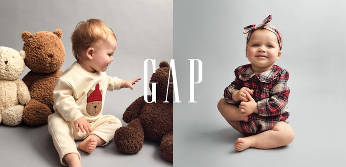 To spædbørn sidder med plysbamsebamser. Den ene har en cremefarvet dragt med et bamsemotiv; den anden er iført en rød og ternet dragt med pandebånd.