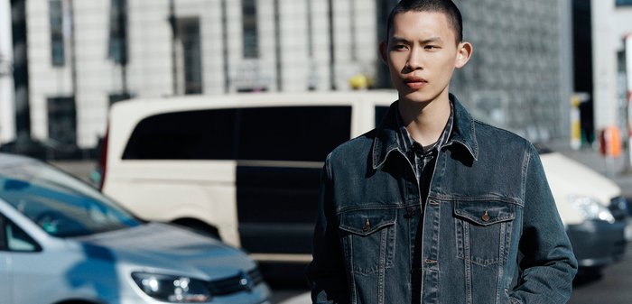 Denim jacket with two chest pockets and a collared neck, worn over a checkered shirt. The fabric is dark blue with a textured appearance.