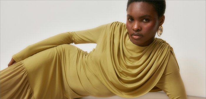 Mujer reclinada, vestida con un vestido drapeado color mostaza y grandes pendientes de aro dorados, fondo neutro, mirada directa a la cámara.