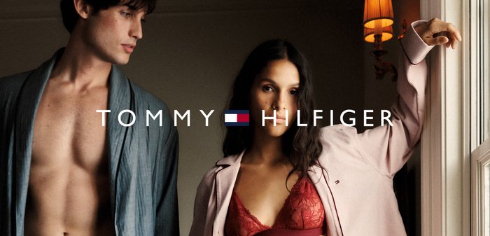 Zwei Frauenmodelle tragen entspannte Nachtbekleidung. Das männliche Modell trägt einen blauen Morgenmantel, und das weibliche Modell hat eine rosa Jacke über einem roten Spitzenoberteil an. Markenname angezeigt.